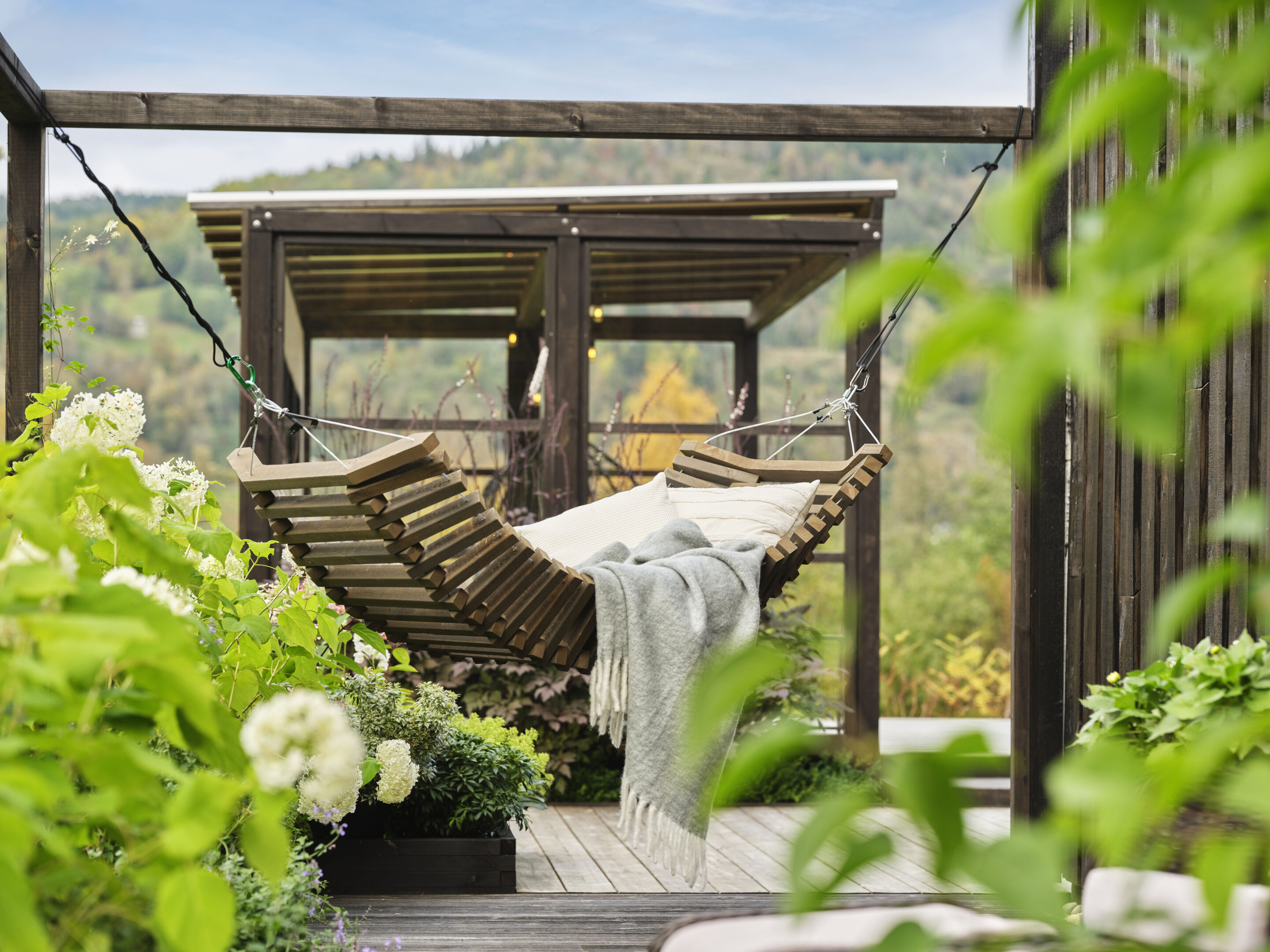 Foto fra Marte Helgetun sin hage, Overbygd pergola i MøreRoyal svart i bakgrunnen. Hengekøye i MøreRoyal grå i forkant. I hengekøyen ligger det myke puter og pledd. Frodige blomster og planter på venstre side av hengekøyen.